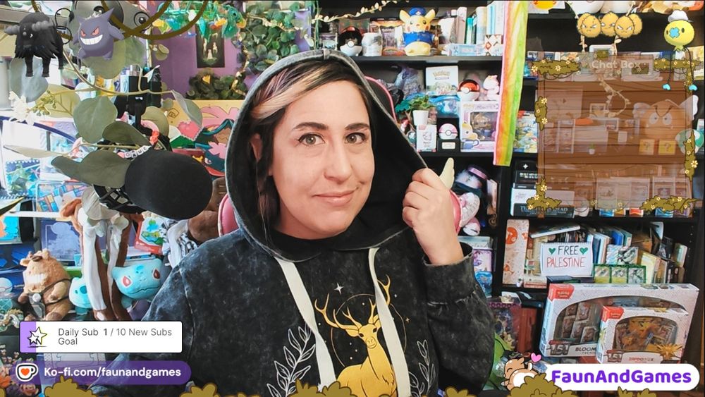 nonbinary streamer FaunAndGames a femme perceived person with tan skin and pink and black hair pulls back the hood on their cloak to smile at the camera.