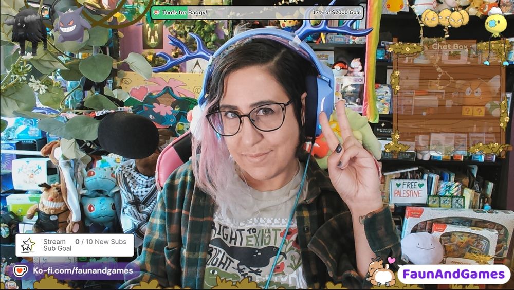 Nonbinary streamer FaunAndGames, a tan skin femme perceived person with brown, blue, and purple hair, is wearing gaming headphones with antlers on them. They are smiling at the camera and making a peace sign. 