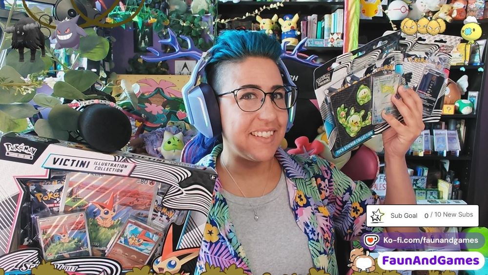 Nonbinary streamer FaunAndGames, a tan skin femme perceived person with brown, blue, and purple hair, is wearing gaming headphones with antlers on them. They are holding up a bunch of new Black and White Pokemon card boxes and smiling at the camera. Around them is a Twitch  stream setup featuring and a whole bunch of Pokemon, Nintendo,  and nerdy memorabilia.