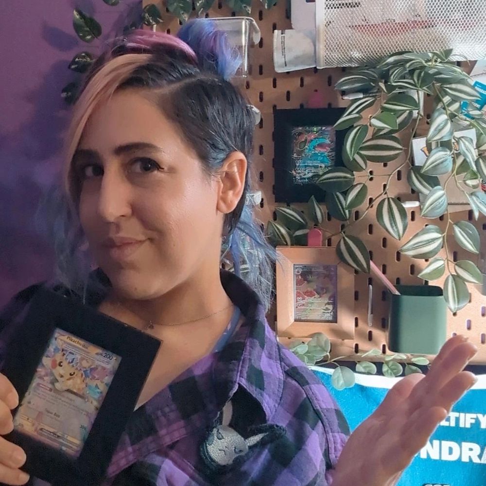 Similar to the first picture, nonbinary streamer FaunAndGames with pink, blue, and dark brown hair is wearing a purple and black checkered shirt. They are holding up a fancy Pikachu Pokémon card in a nice black frame towards the camera and smiling but are turned away and looking more cheeky. They are showing off a plant covered board behind them with more Pokemon TCG cards in frames.