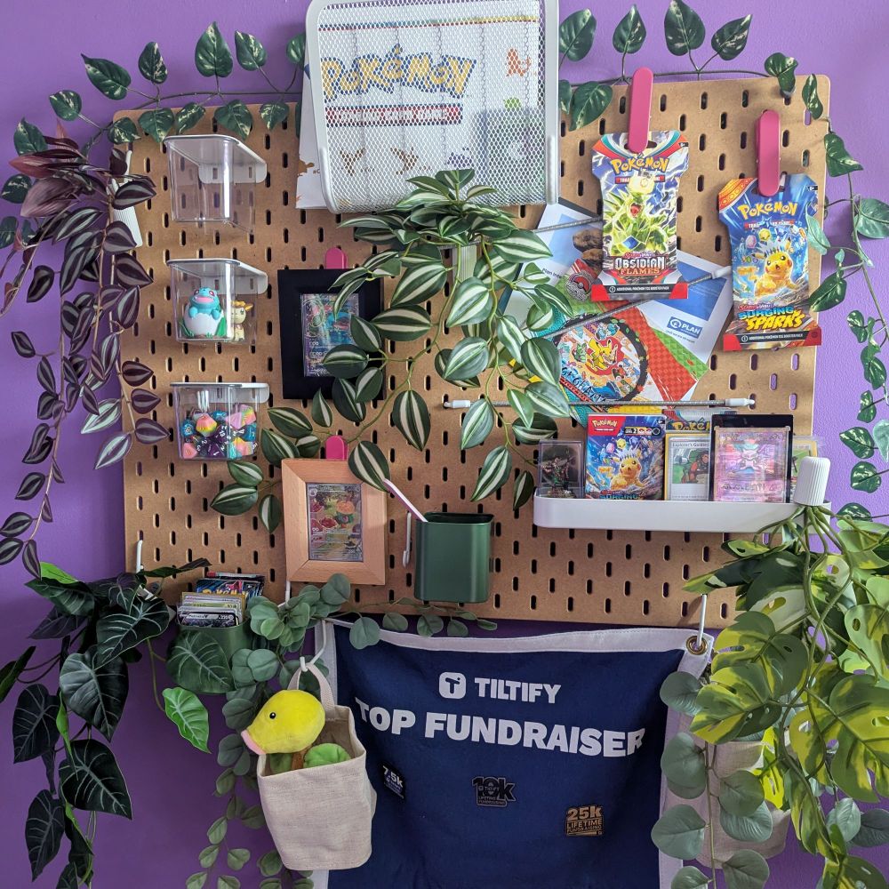 A zoomed out picture of the same plant covered wooden board with Pokemon TCG cards in frames. It shows that there are even more nerdy things on the wood board than in the last picture. Like a Pokemon plushie, figurines, colourful dice, and more unopened Pokemon card packs.