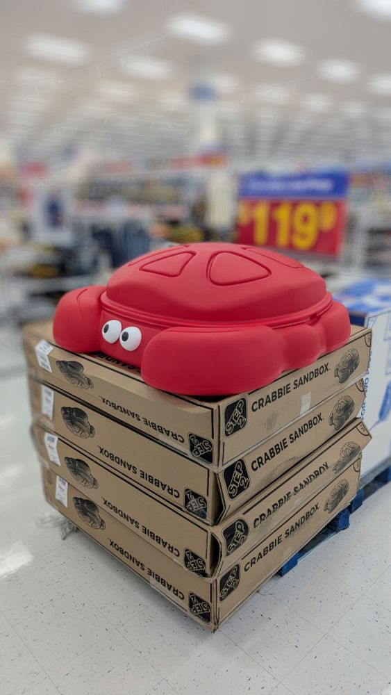 A red crab shaped sandbox for kids sits on top of a stack of 4 large cardboard boxes that read "Step 2: Crabbie Sandbox"