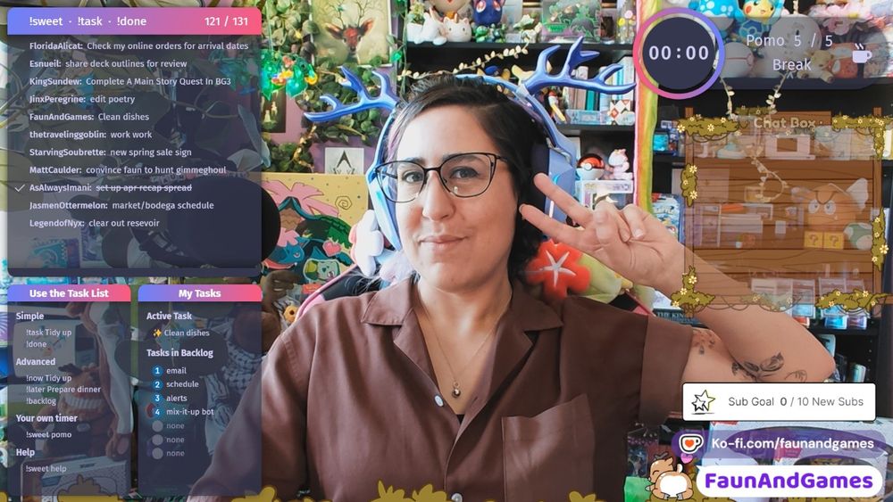 Nonbinary streamer FaunAndGames, a tan skin femme perceived person with brown, blue, and purple hair, is wearing gaming headphones with antlers on them. They are smiling at the camera and making a peace sign. Around them is a Twitch coworking stream setup featuring Super Sweet Bot. Shows a task list and other co-working applications.