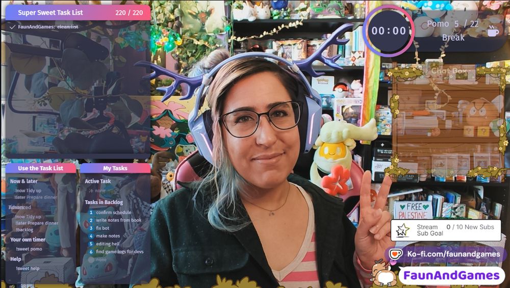 Nonbinary streamer FaunAndGames, a tan skin femme perceived person with brown, blue, and pink hair, is wearing gaming headphones with antlers on them. They are making a peace sign at the camera with a Twitch coworking stream setup around them. 