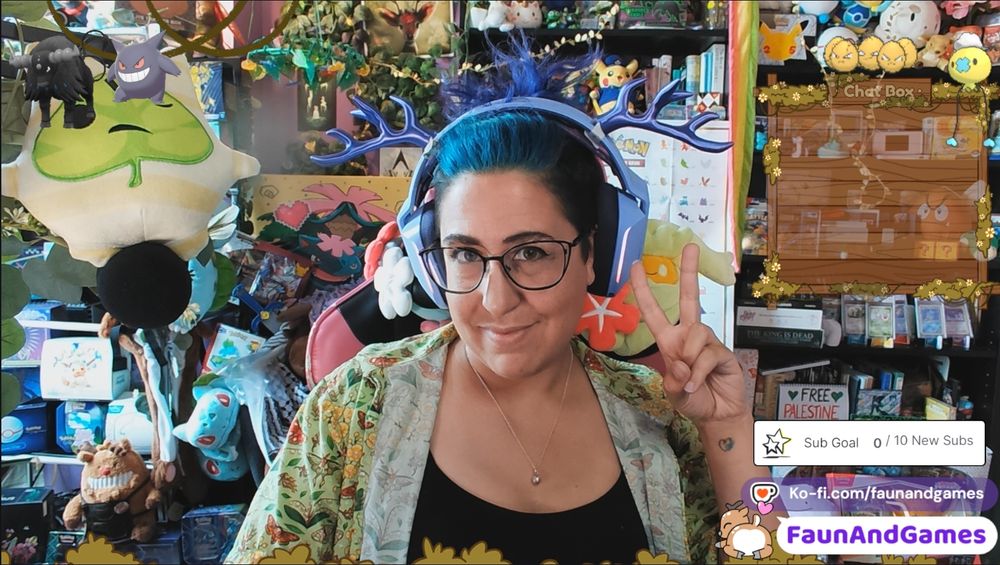 Nonbinary streamer FaunAndGames, a tan skin femme perceived person with brown, blue, and purple hair, is wearing gaming headphones with antlers on them. They are smiling at the camera and making a peace sign.