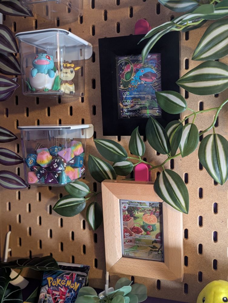 A zoomed in picture of a plant covered wooden board with Pokemon TCG cards in frames. There are also other nerdy things on the wood board like a Pokemon plushie, figurines, and colourful dice.