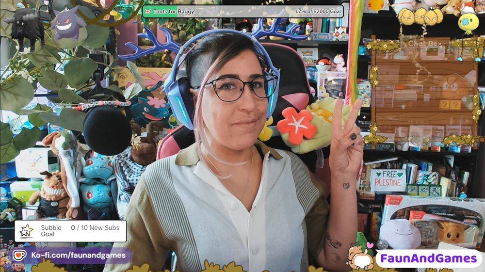 Nonbinary streamer FaunAndGames, a tan skin femme perceived person with brown, blue, and purple hair and partly shaved head, is wearing gaming headphones with antlers on them. They are smiling at the camera and making a peace sign. 