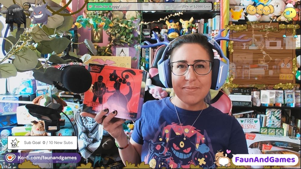 Nonbinary streamer FaunAndGames, a tan skin femme perceived person with brown, blue, and purple hair and partly shaved head, is wearing gaming headphones with antlers on them. They are smiling at the camera and holding up a Pokemon TCG card box. Around them is all sorts of Pokemon collections and nerdy stuff.