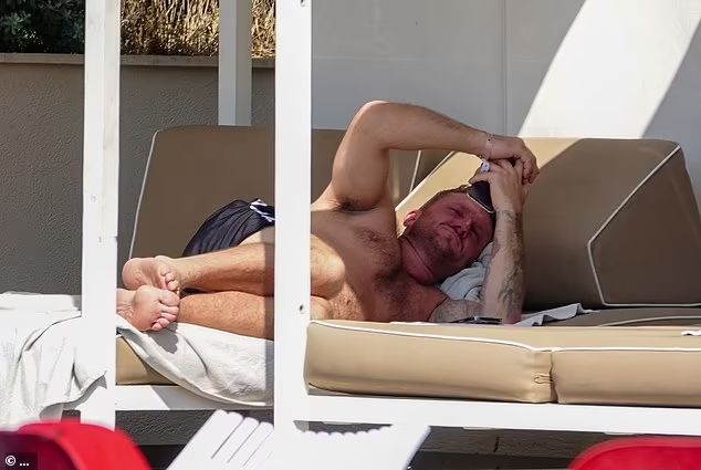 An image of notorious fascist Stephen Yaxley-Lennon (who styles himself as 'Tommy Robinson') loafing around at a resort in Cyprus during the height of far right riots in 2024 and while avoiding a warrant for his arrest in the UK, after he was found guilty of libeling a 15 year old Syrian refugee and refused to present himself in court. His face is an unpleasant shade of red and he is squinting up towards his mobile phone. It is speculated that at this moment he is inciting further racist violence on social media.