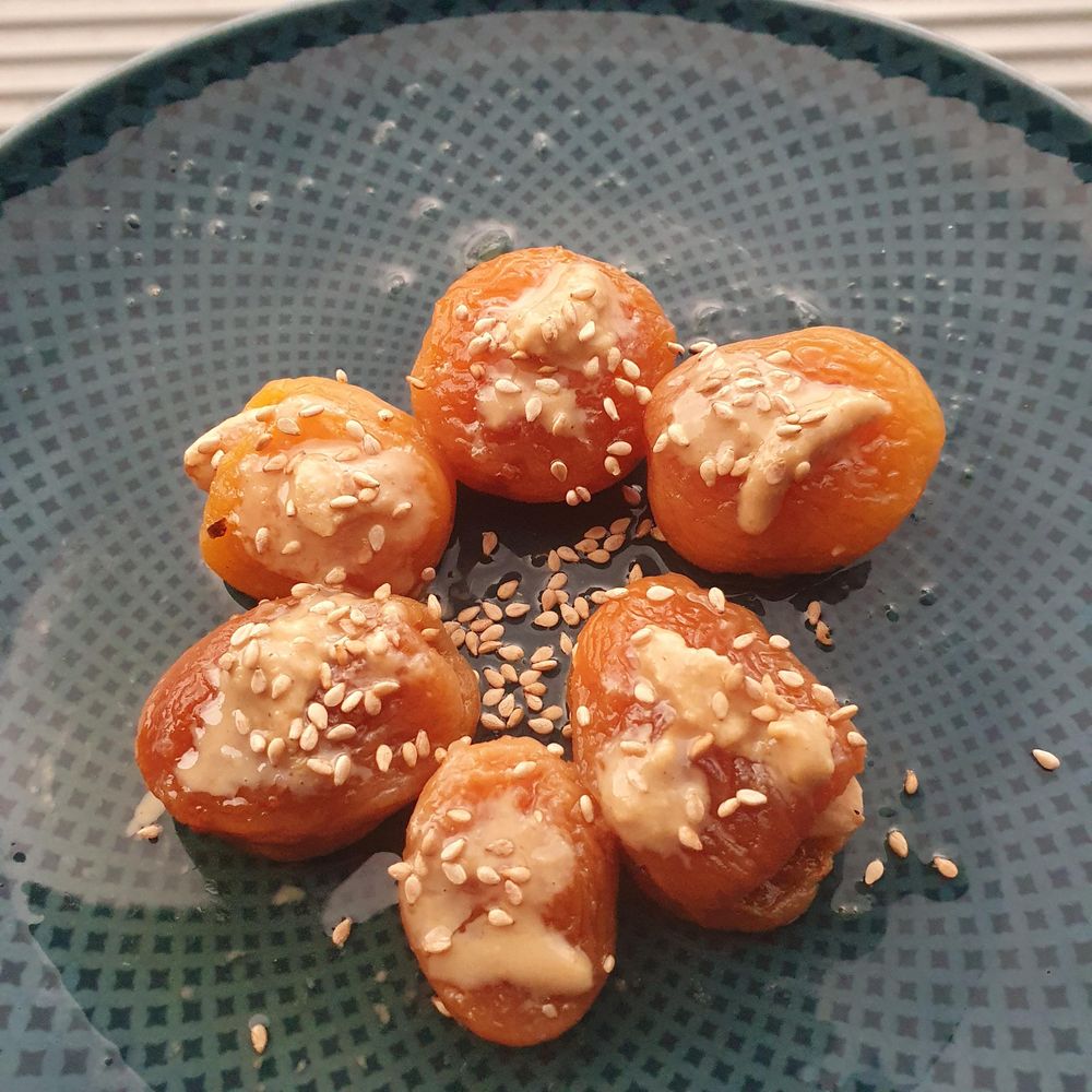 6 dried stewed, filled apricots arranged on a plate, with tahini drops and sesame seeds on top. 
