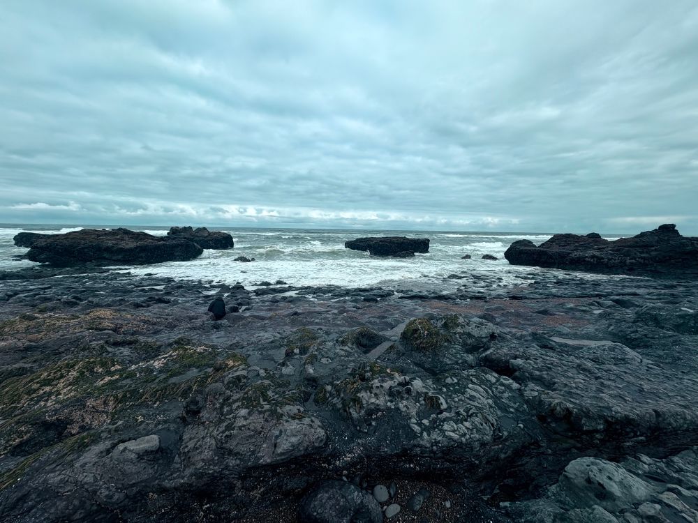 Photo of the beach in central coastal Oregon. It was dark and overcast in the morning. There is an outcropping of rock in the foreground and three large rocks in the background that have ocean waves breaking on them. 