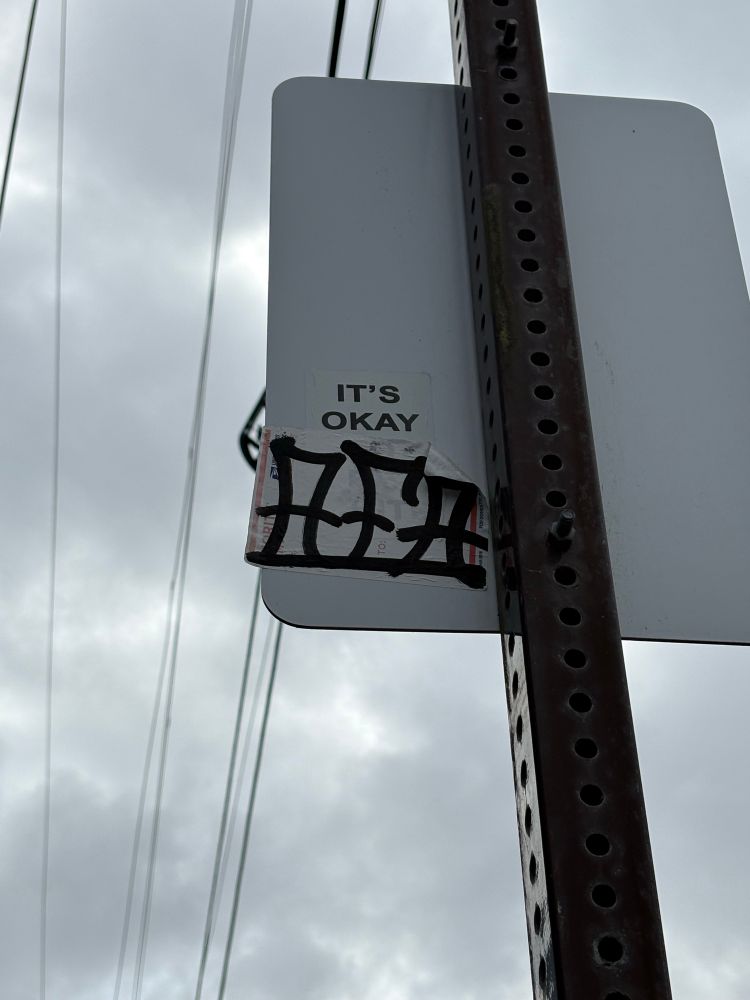 A back of a street sign with two stickers on it. The first sticker says “it’s okay” and the second sticker says “AFA” to make a silly little rhyme “it’s okay, AFA” 
