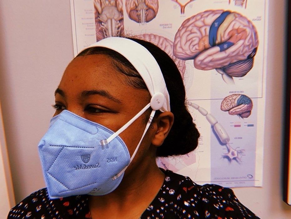 Selfie of a Black woman in a healthcare setting. She is wearing work scrubs and a mask and there is a poster of the human brain and neurons are shown in the background. 