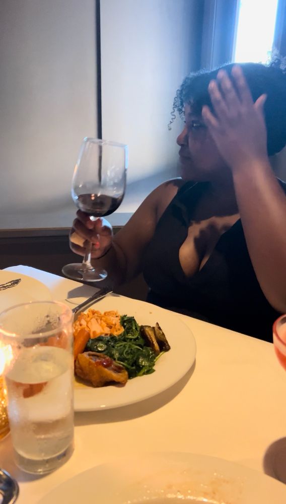 Beautiful Black woman sitting in a restaurant looking away from the camera. She is wearing a Black dress and holding a glass of wine. She appears to being eating, in more ways than one. 