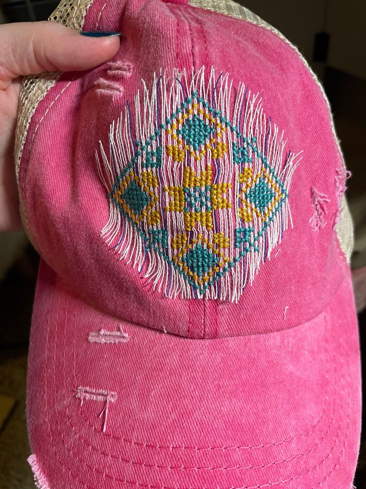 Traditional Palestinian cross stitch (tatreez) in teal and gold on a pink hat. It has been stitched over a grid of white thread called waste canvas. Half of the waste threads have been pulled out to demonstrate the process.