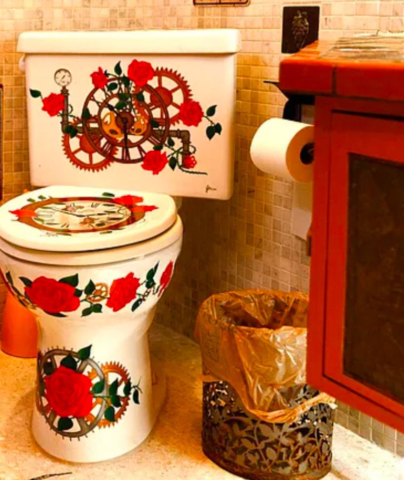 a photo of a bathroom with an extremely ornately painted toilet (white with roses, gears and clocks), a red sink, and between them, a metal oval-shaped trash made of leaf/plant cutouts, with a plastic shopping bag as the trash bag