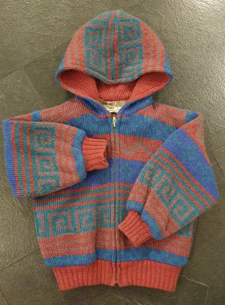 En strikka ulljakke i rosa, grønn, blå og lilla, med hette. Hetta har rosa strikka fôr, jakken har hvitt bomullsfôr.