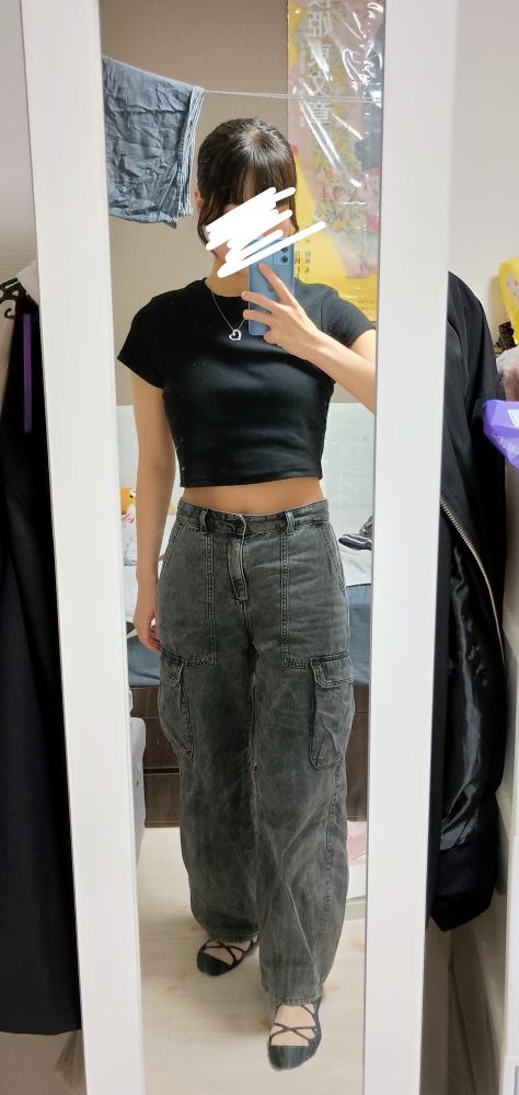 黒のミニT、グレーのジーンズ、黒のレースアップの靴下、胸には銀のネックレス、の女性が青いスマホを使って鏡越しに自撮りしている