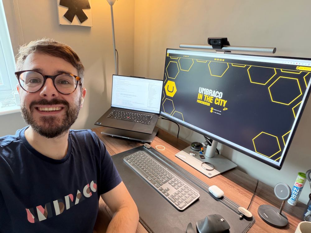 A selfie of Rick with a computer desk, laptop, large monitor, keyboard and mouse in the background. The computer screen displays the Umbraco in the City logo and website.
