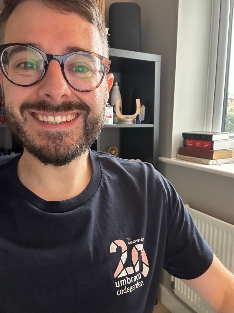 A selfie of Rick, who has dark hair, a dark beard and glasses. He is wearing a navy tshirt with a large “20” print on it and “Umbraco Codegarden” underneath. The background is an office room with a shelving unit and a window.
