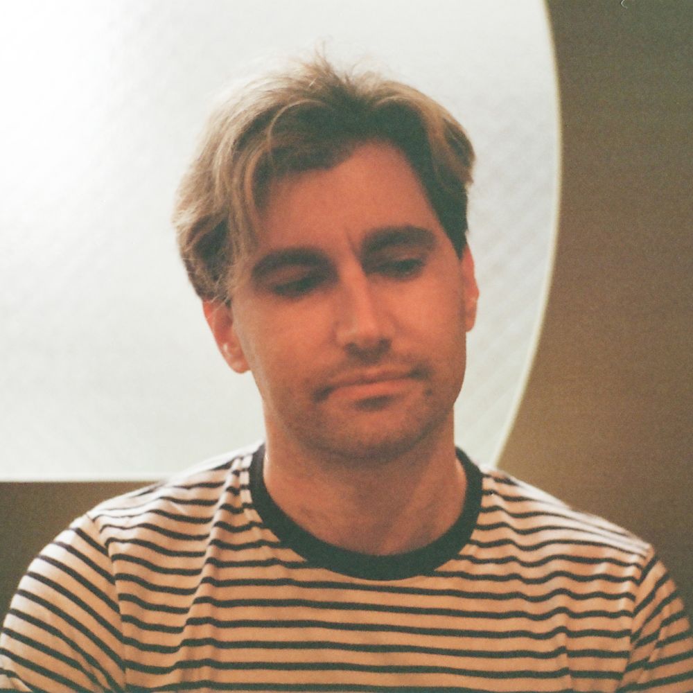 A photo of VESSITT, he is looking down and to the left and wearing a black and white stripped shirt.  There is a blurry window in the background backlighting him.