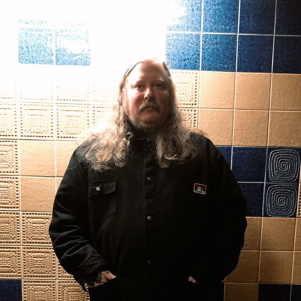 A photo of Rhins, they are center and looking at the camera. They are wearing a black jacket and have long hair below the shoulder. Behind them is a yellow and blue tiled wall.