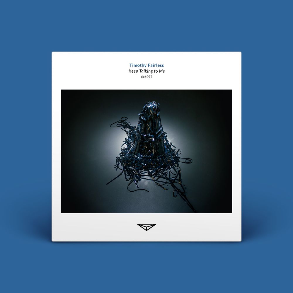 Album cover made to look 3d with a shadow below and behind it sitting in a blue background. The album cover is white with text centered at the top that reads “Timothy Fairless, Keep Talking to Me, de6073.” The center features a photograph of magnetic audio tape in a large pile on the floor, the center juts up to an almost unnatural point and the edges are frayed out in all directions.  It is lit from above so it looks like a spotlight is on it which makes the tape shimmer. The bottom feature the triangular Dragon's Eye Recordings logo. 