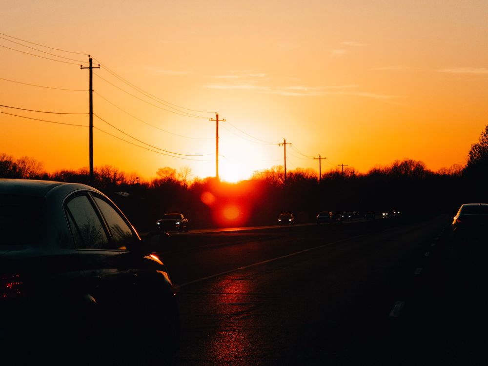 A vibrant sunset over a highway with cars driving in both directions. The sky is painted in warm hues of orange and yellow, casting a golden glow on the road. Silhouettes of trees and utility poles line the horizon.
