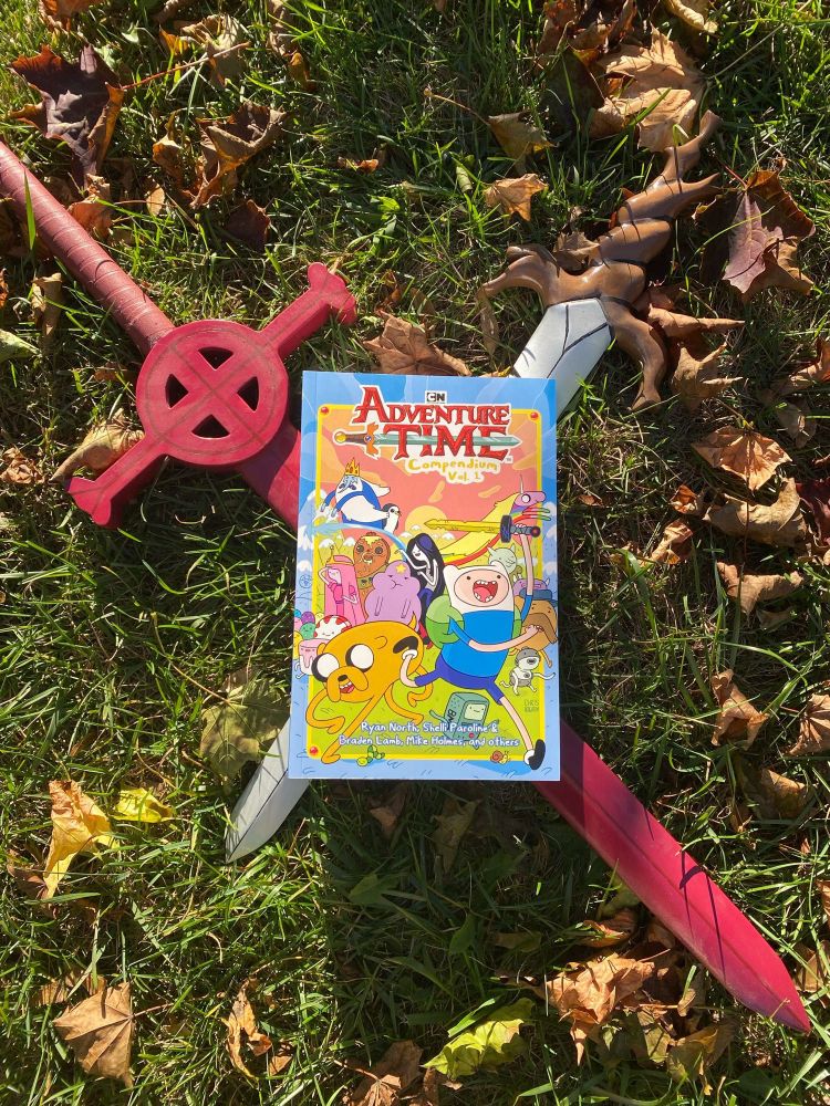 A huge beautiful compendium of the Adventure Time Comics lying on two crossed replica swords from the cartoon - the demon blood and root swords. It's on a grassy field with autumn leaves in some golden hour light, looking very purchaseable