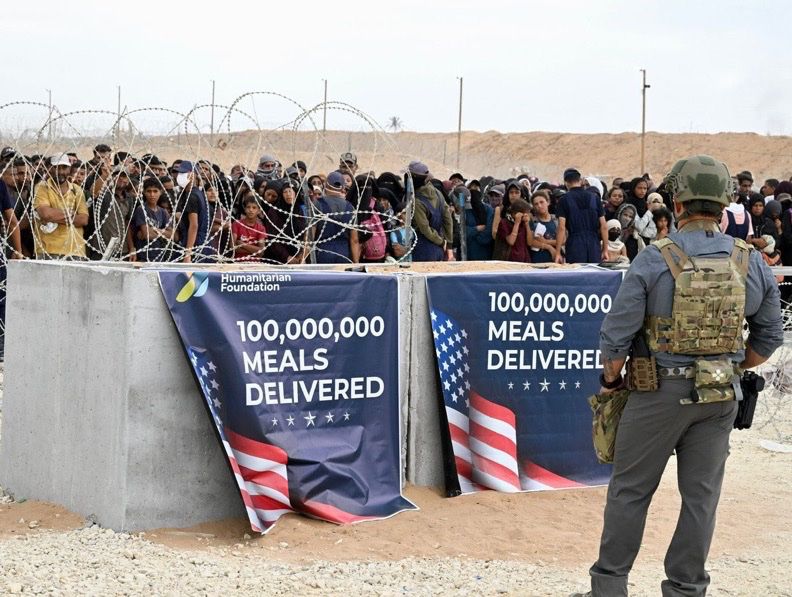 Two posters are seen draped from barbed wire-covered concrete. The posters say 100,000,000 meals delivered with a flying US flag. In the background are people awaiting food aid and in the foreground is a soldier.