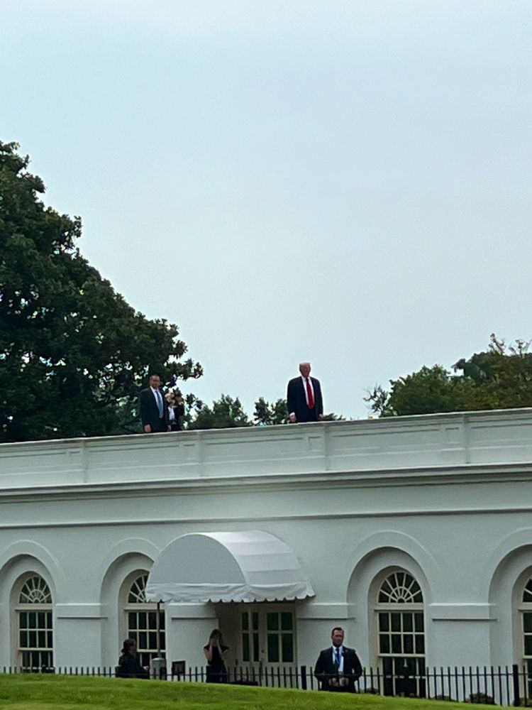The roof of the briefing room is seen with Trump in his suit standing still on it. A secret service agent is seen behind him and another on the ground below.