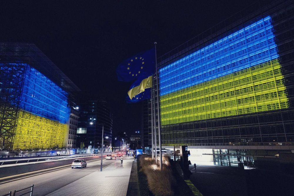 A night time photo of the The European Commission buildings in Brussels. The buildings on either side of the main road leading to Schuman are illuminated in the blue and yellow colours