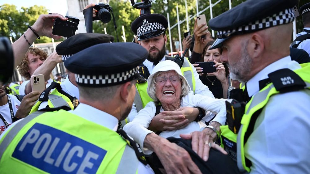 Four Met police officers are seen carrying the 89 year old protester away. Her face looks strained with anguish, her eyes closed as she is taken away. Others around are taking photos. Credit to Chris J Ratcliffe/AFP.