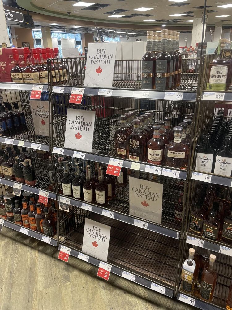 A picture of a liquor store in British Columbia. Gaps in the shelves where US spirits would be are occupied by signs which say BUY CANADIAN INSTEAD. Photo credit to Riley Donovan