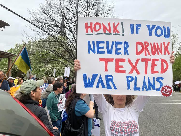 A woman at a #HandsOff protest holds up a sign which reads “HONK IF YOU NEVER DRUNK TEXTED WAR PLANS”