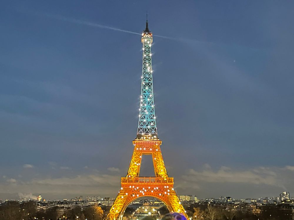 The Eiffel Tower in Paris at night with bottom half in yellow and top half in blue with the spotlight at the top shining across the city