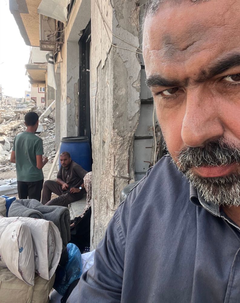 A picture of Motasem posted by him at the same time as the messages. He is standing outside a building with rubble and two other men in the background. 
