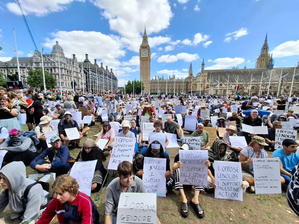 Hundreds sit on the ground in Parliament Square with Big Ben in the background. They are holding placards which read “I oppose genocide. I support Palestine Action”