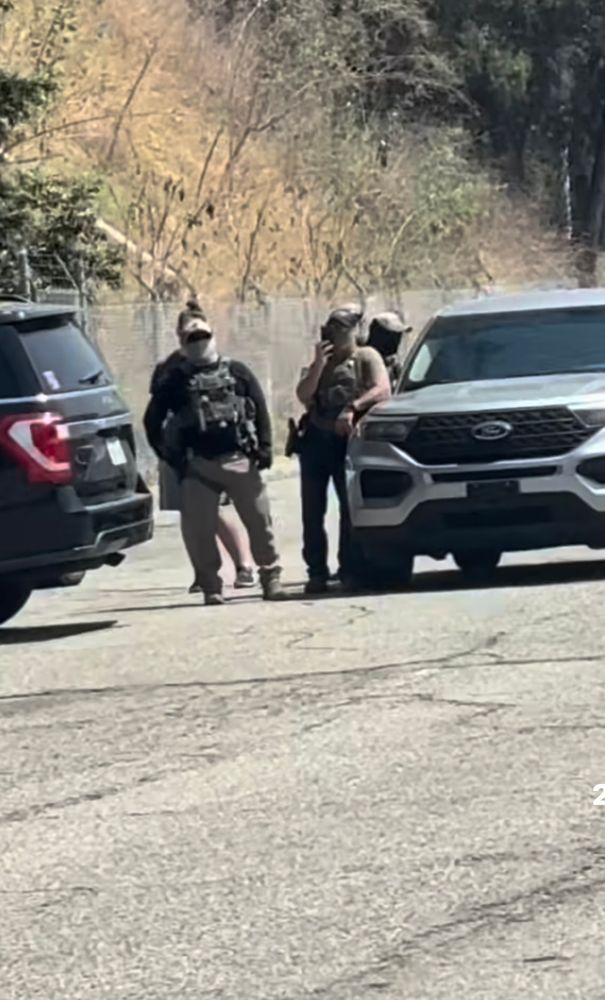Two masked men are seen resting on their cars outside Dodgers stadium car park. Another two people are seen behind them. One is on the phone