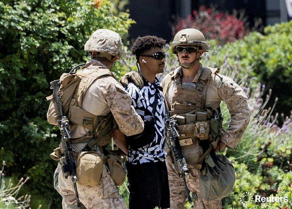 Two Marines in army combat outfits and guns are seen detaining a young black man in a black and white top, wearing sunglasses with air pods in his ears.