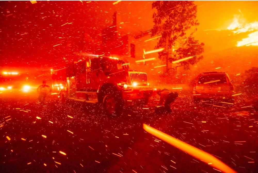 A fire truck is engulfed in parks and flames are seen on the hill. The entire photo is red and orange from the fire