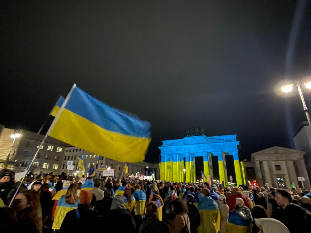 Demonstrators seen gathered in Berlin at nighttime. In the foreground is a Ukraine flag, in the background is the Brandenburg Gate in the yellow and blue colours