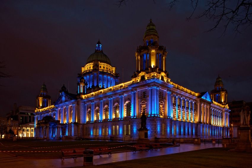 Belfast City Hall in Northern Ireland is lit in blue and yellow against the dark night sky
