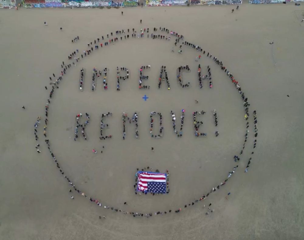 People form a large circle and the words IMPEACH + REMOVE. There is also an upside down star and stripes, a historical sign of distress. The photos is taken directly above looking down on the people and words.