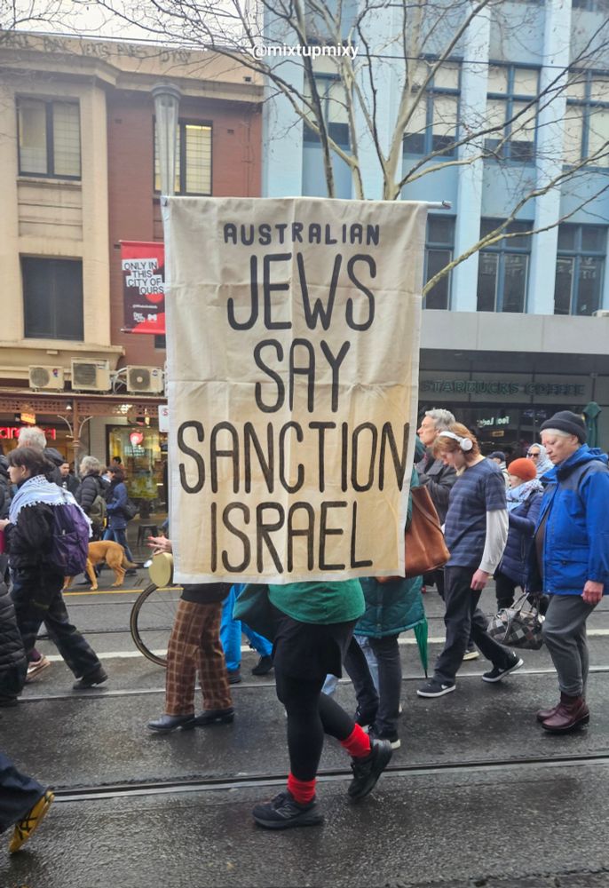 A lady is walking along the street holding a large banner which says;
Australian Jews Say Sanction Israel.