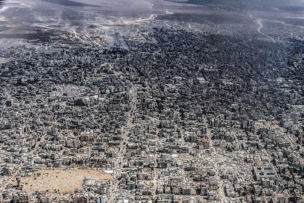 From above everything looks grey or black. Smoke is seen rising from building ga at the top of the photo. In the foreground many buildings have collapsed entirely. Photo credit to Heidi Levine taken for the Washington Post.