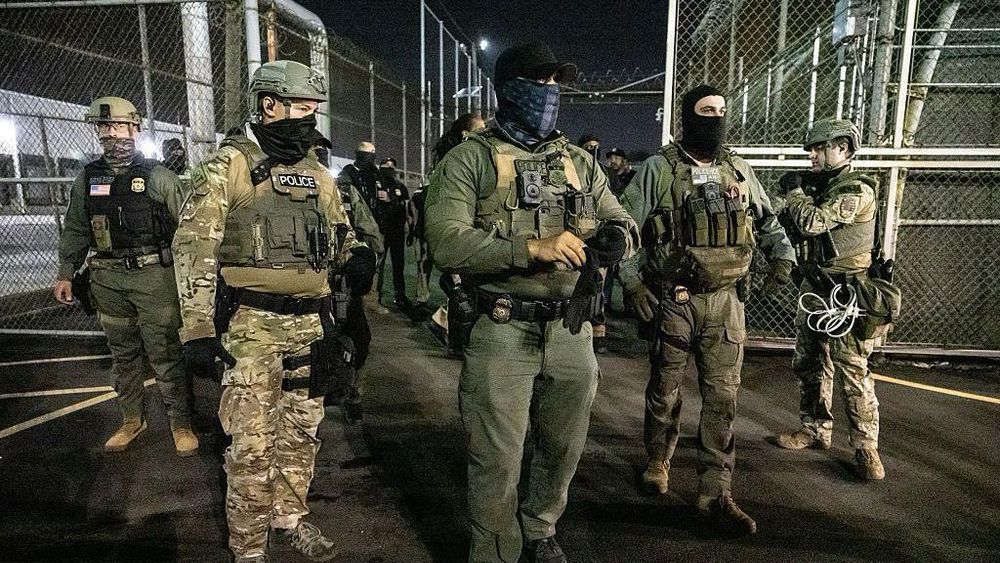 A night shot of multiple ICE agents masked as always outside a temporary private prison.