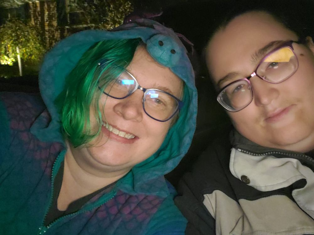 On right, a human with dark hair and purple glasses sits in an 80s looking jacket and a half-smile. Opposite them on the left is a beautiful grinning emerald haired woman wearing blue glasses and a dinosaur onesie.