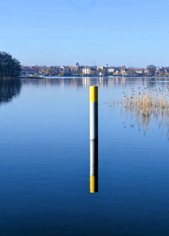 Stiller See, am Ufer gegenüber Häuser. Im Vordergrund ein grau-gelber Pfosten, der sich im Wasser spiegelt.