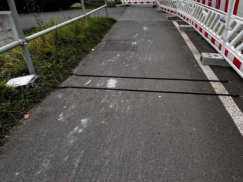 Asphaltierter Fußweg und jede Menge Absperrgitter. Über den Fußweg laufen zwei parallele Kabel, die aus einem Plastikkasten kommen.